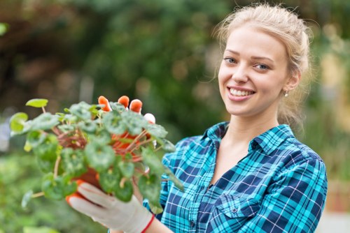 A young woman with blonde hair tied back, wearing a blue and green checkered shirt and gardening gloves, is smiling while holding a small potted plant with green leaves and some reddish-pink flowers. The background features a blurred outdoor garden scene with green foliage, suggesting a well-maintained backyard or front garden area in Parsons Green. The garden surface includes lush green grass and possibly a paved walkway or patio area, with natural daylight illuminating the scene under clear weather conditions. This image exemplifies outdoor gardening activities that might be part of a professional landscaping or hedge trimming service in Parsons Green, emphasizing garden care and plant maintenance.